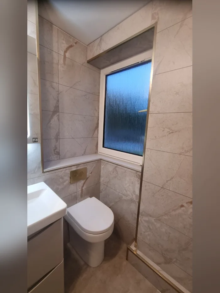 Wall-hung WC with a brass flush plate, beige marble-effect tiles and brass trim round the window