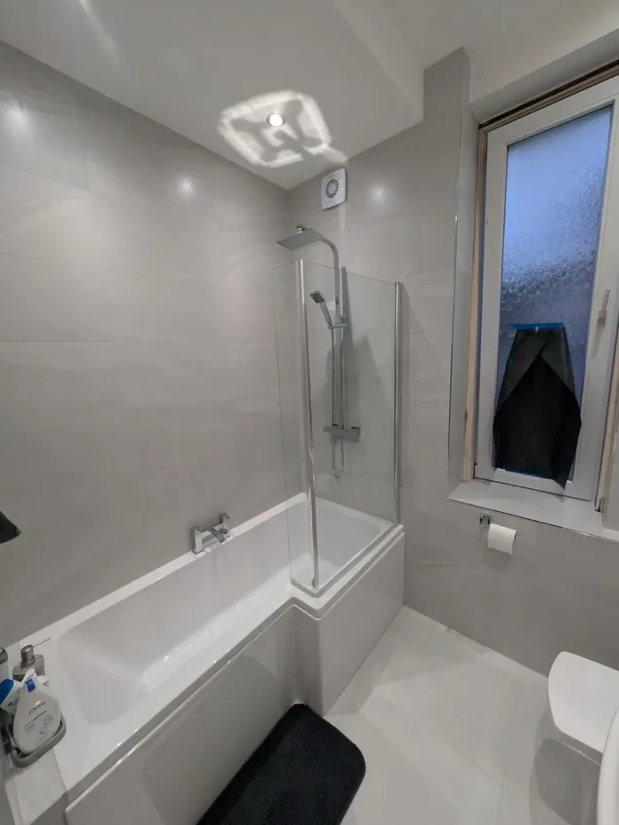 Shower bath with a glass screen and riser shower under a tall frosted window in light grey tiles