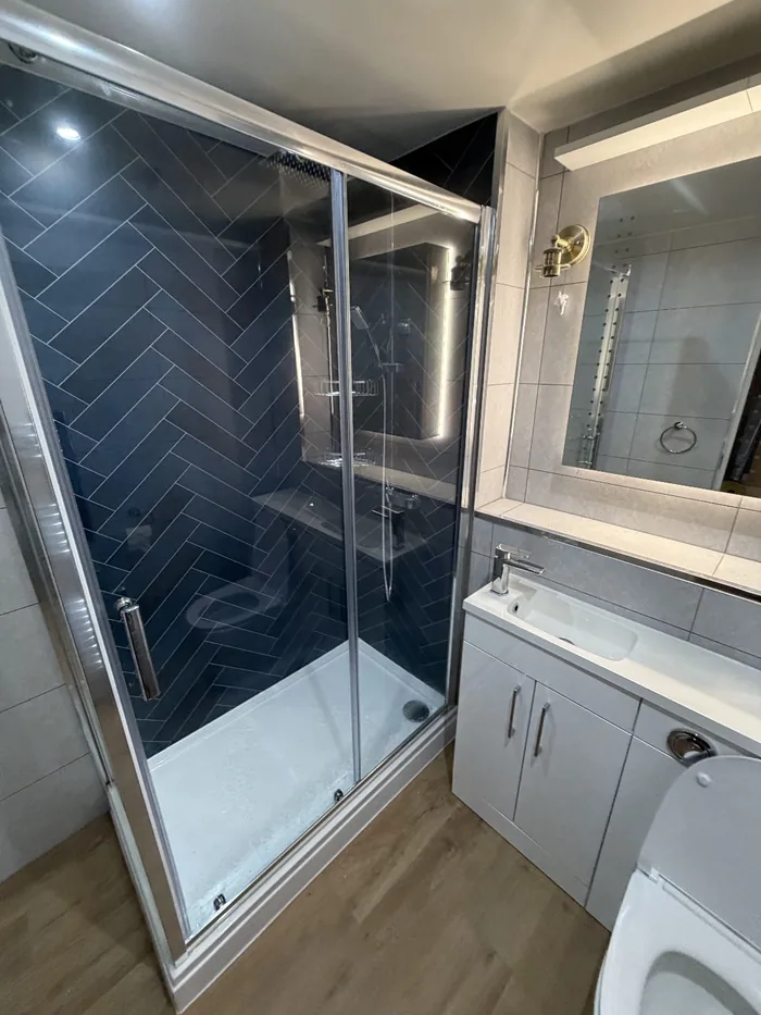 Sliding shower enclosure with navy herringbone tiles, a white vanity and WC unit and a brass wall light