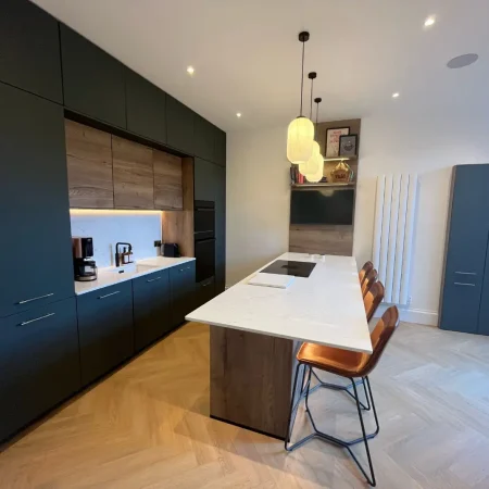 Dark green handleless kitchen with oak-fronted wall cabinets, a white island with tan leather stools and a herringbone floor